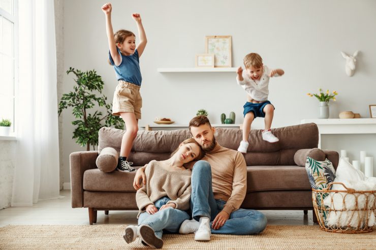 Unhappy parents sitting in front of a couch with kids jumping on the sofa